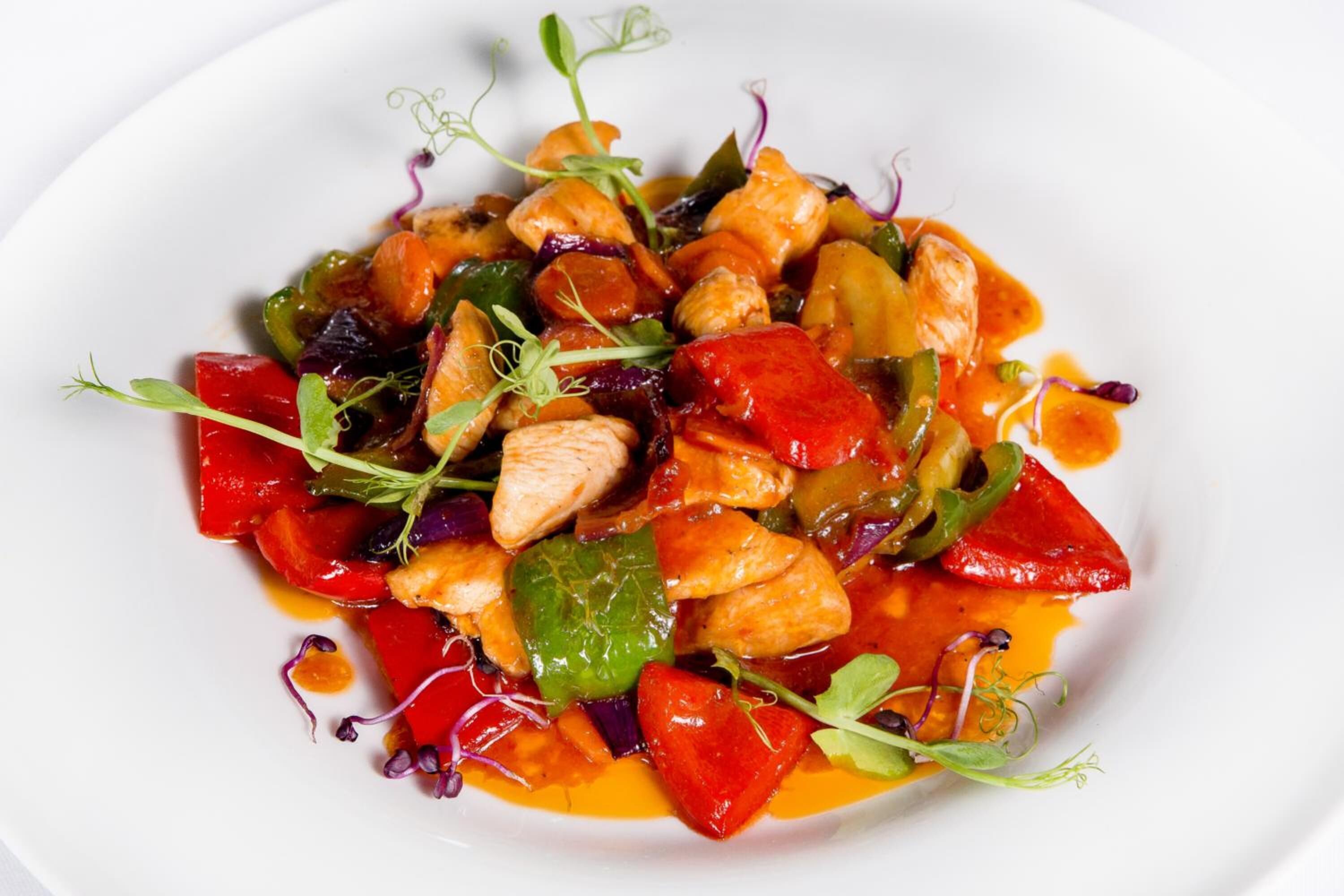 Une assiette blanche contenant un plat coloré composé de légumes et de poulet, agrémenté de jeunes pousses.