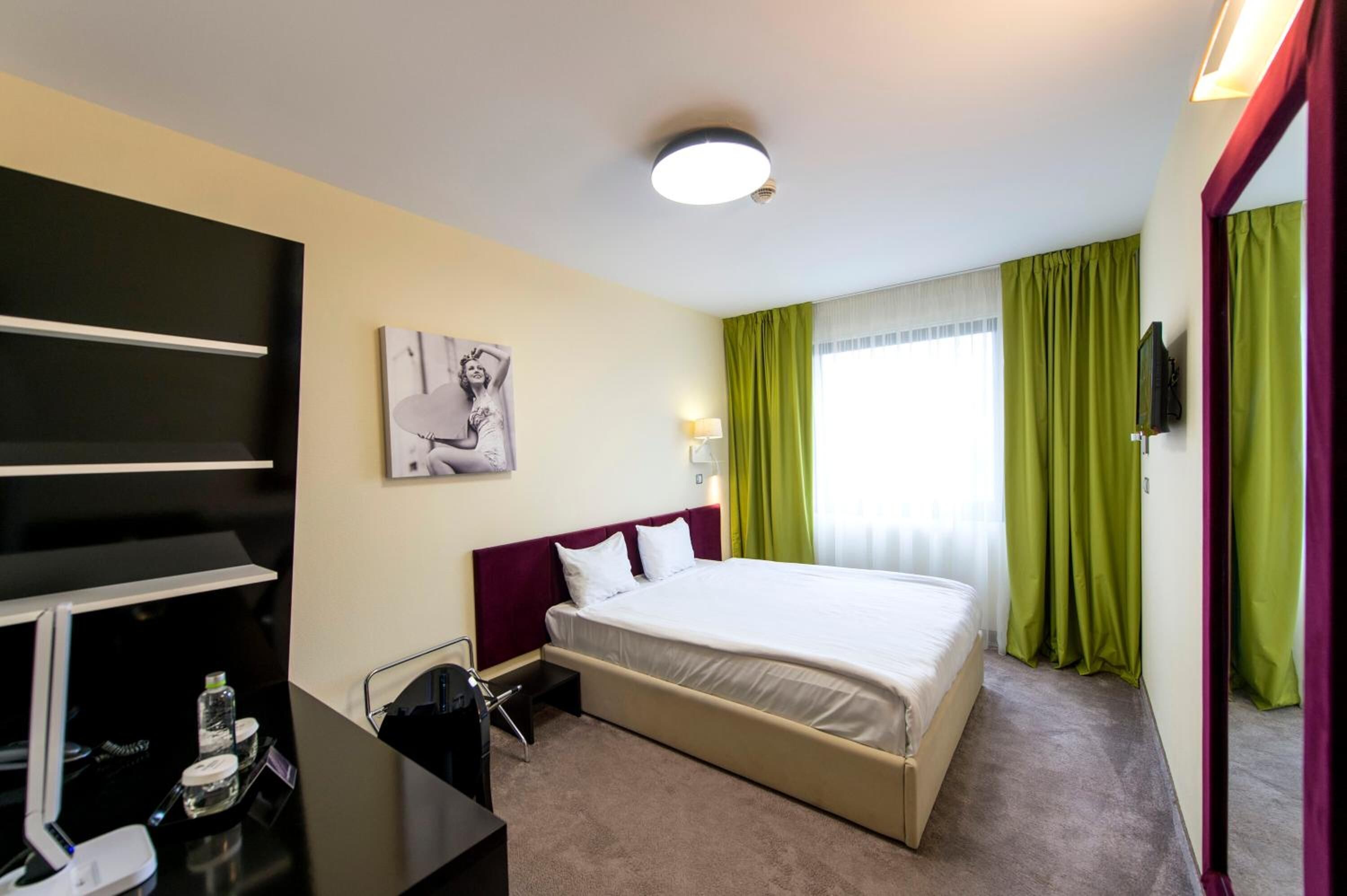 A hotel room with a bed, a desk with a chair, a flat-screen TV, a painting on the wall, and green curtains.