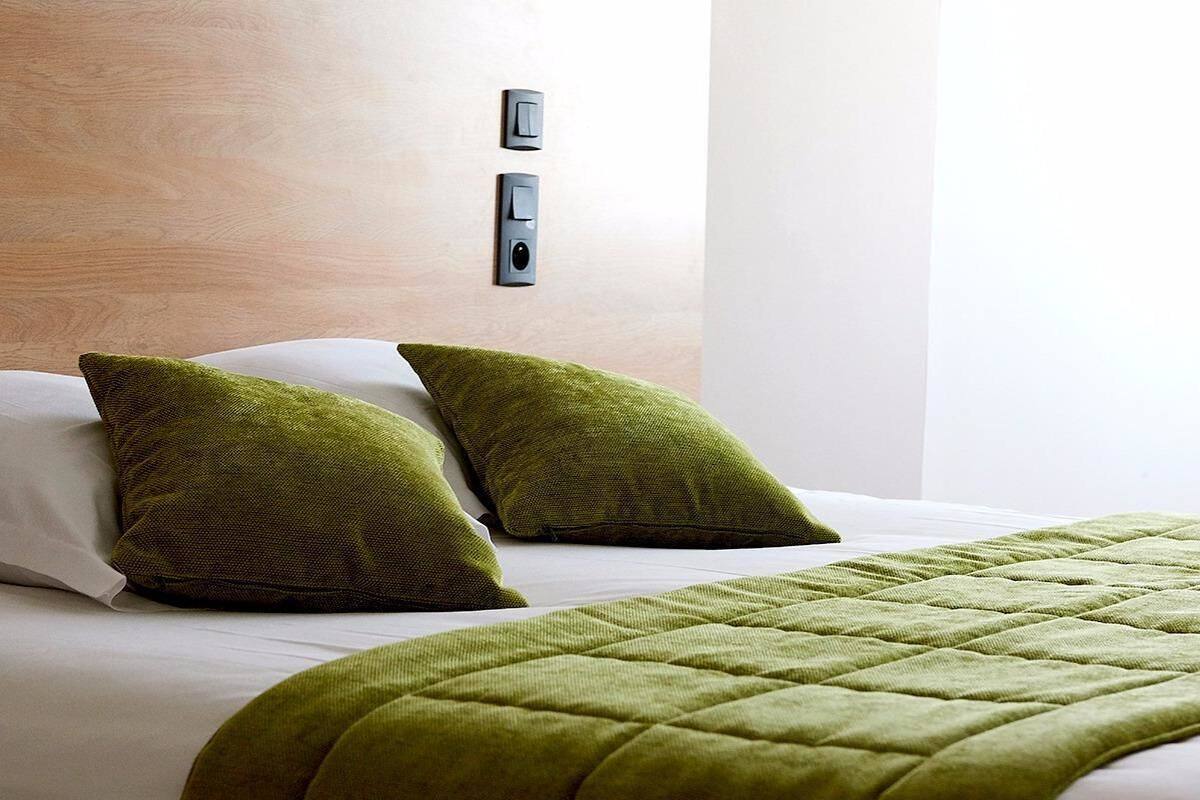 A hotel room with a bed, two green pillows, a quilted green blanket, and a wall-mounted light switch.