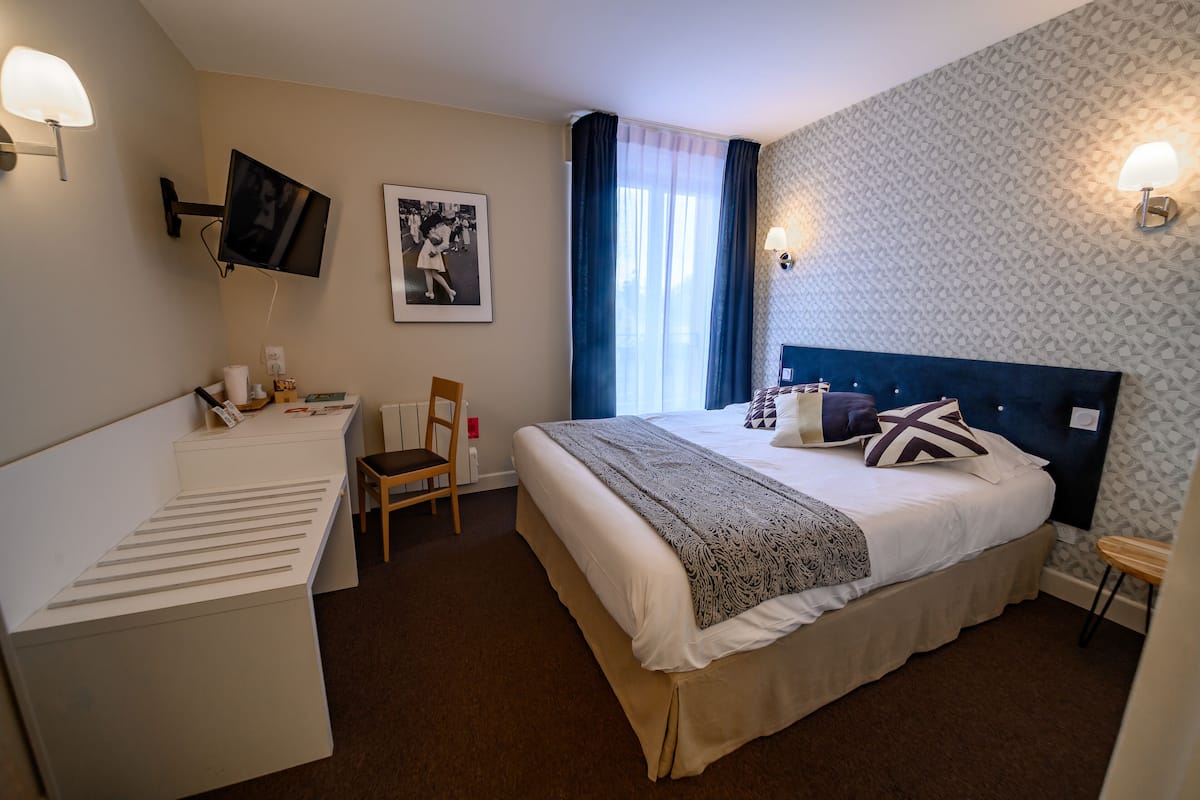 A hotel room with a bed, a desk, a chair, a TV mounted on the wall, and a picture frame.