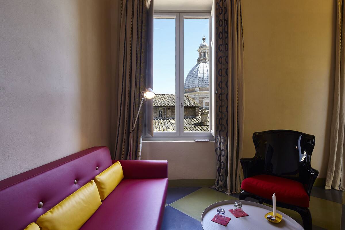 Una habitación con un sofá morado, una silla negra y una mesa con una vela. Una ventana muestra un edificio con una cúpula.