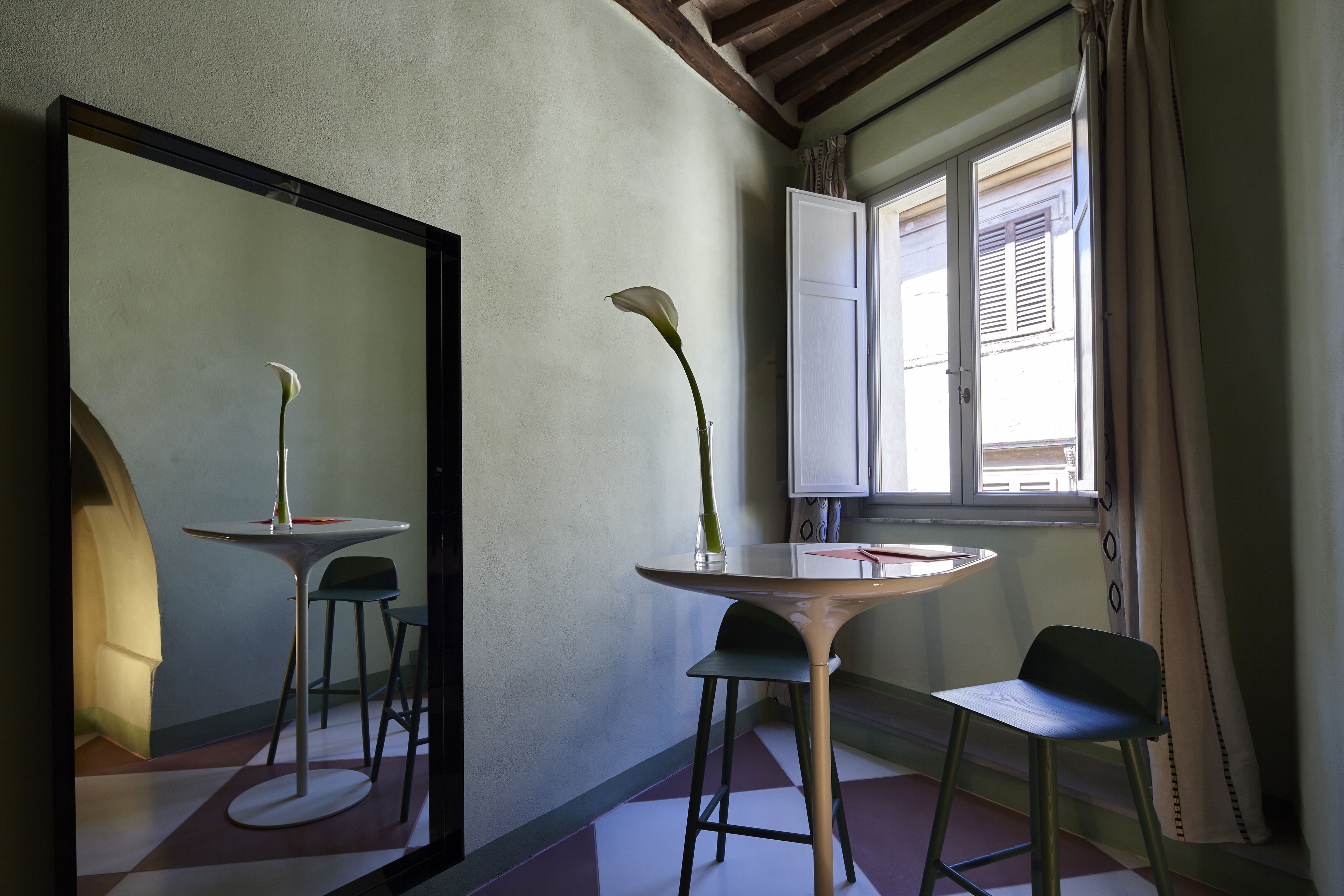 Une pièce avec une table ronde, deux chaises, un vase contenant une seule fleur, un grand miroir et une fenêtre avec des rideaux.