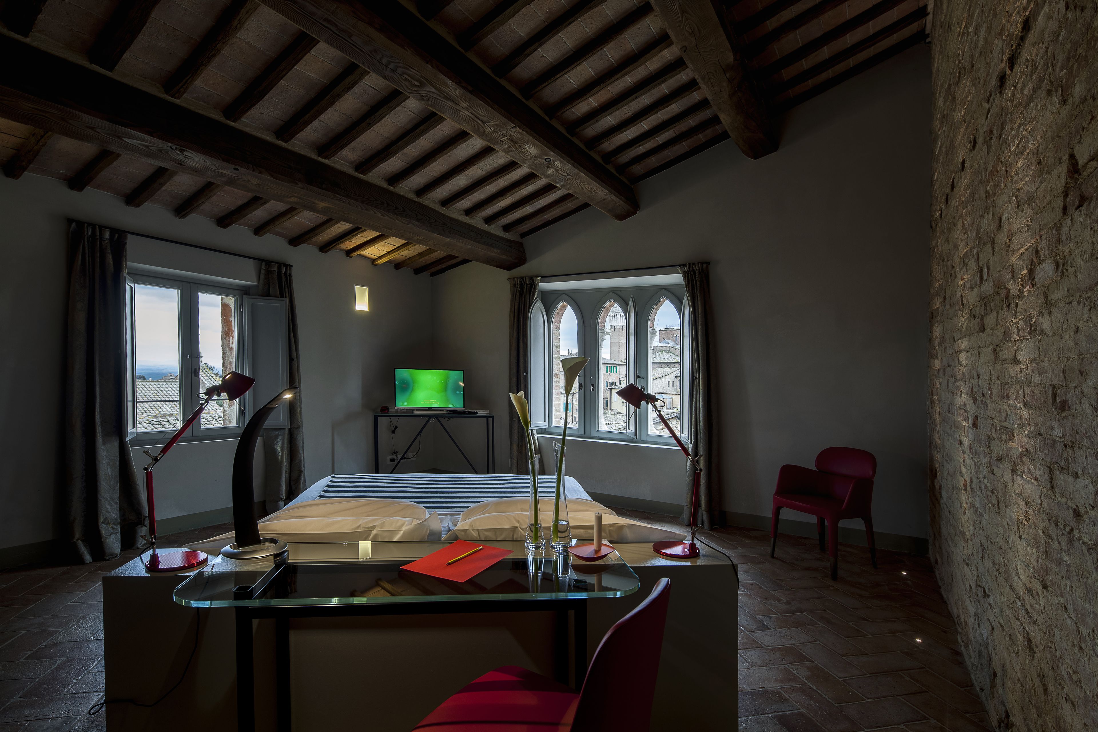 A room with a glass desk, two red chairs, a television, and a view through arched windows.