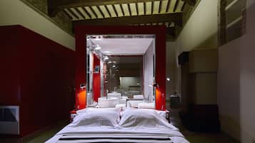 Habitación de hotel con una cama grande, una pared de acento roja, un baño con un espejo grande y un techo con vigas a la vista.