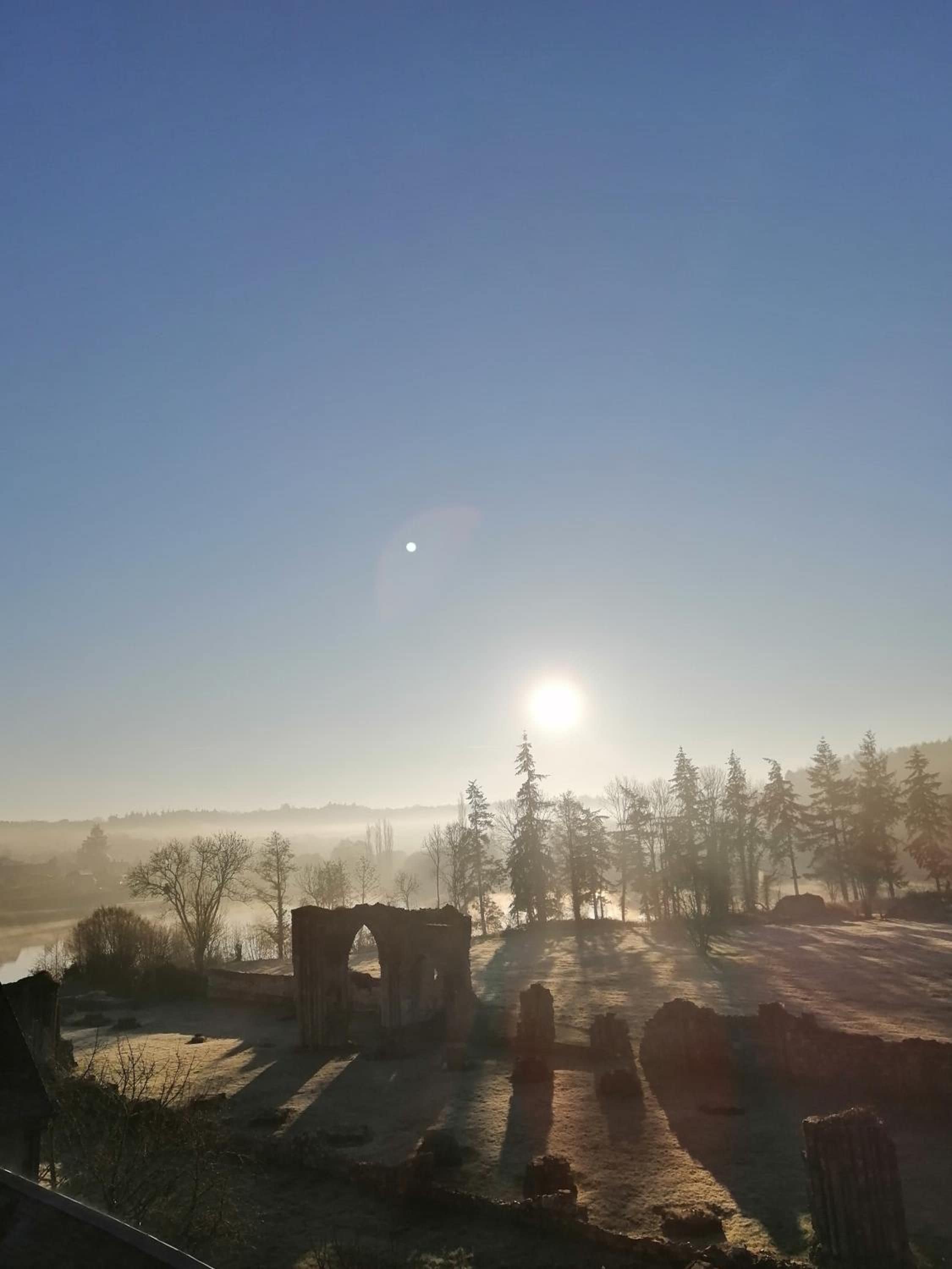 A historical site with ruins and trees, under a clear sky with the sun visible.