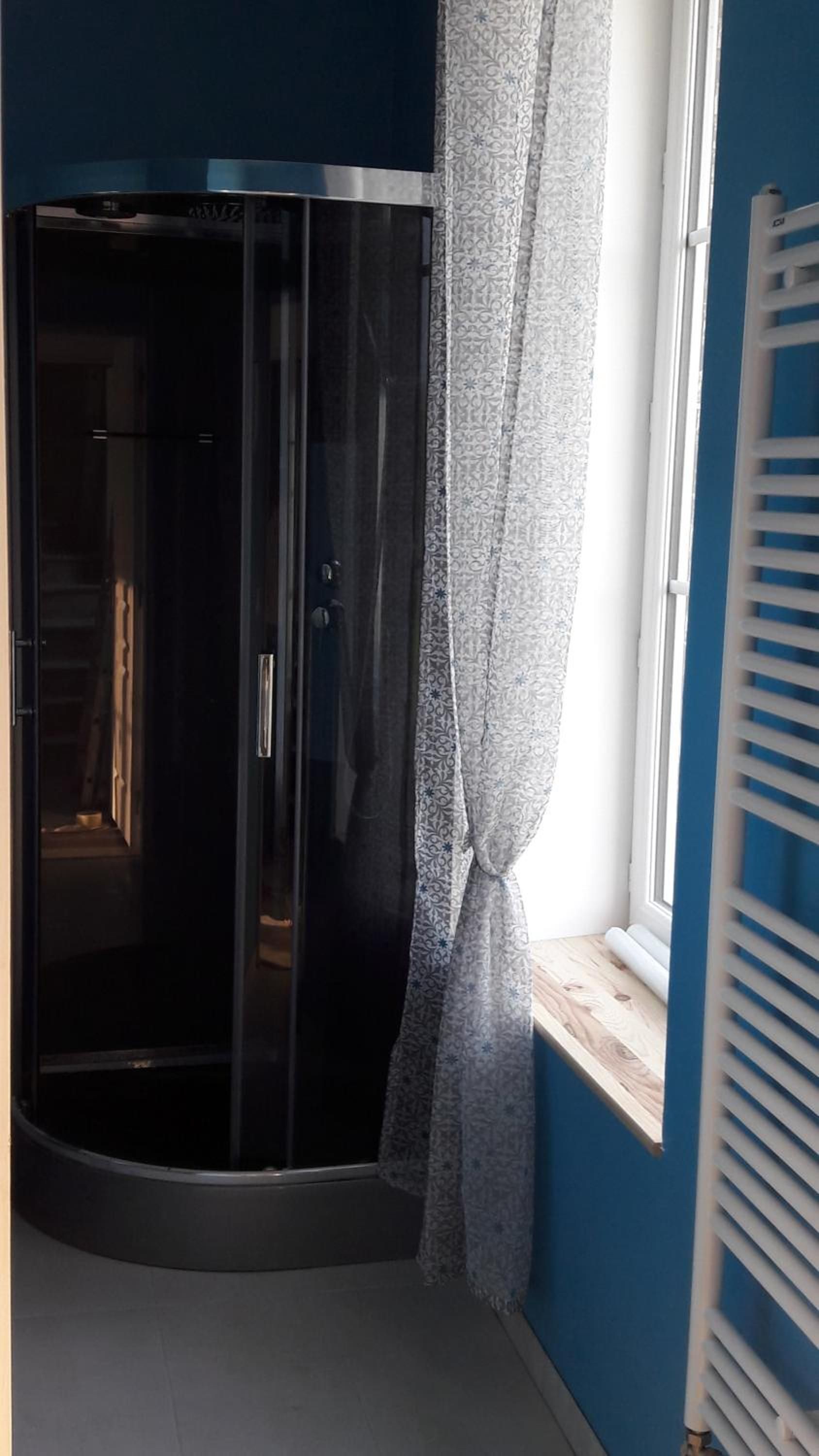 A corner room with a glass shower enclosure, a window with a white radiator, and a patterned curtain.