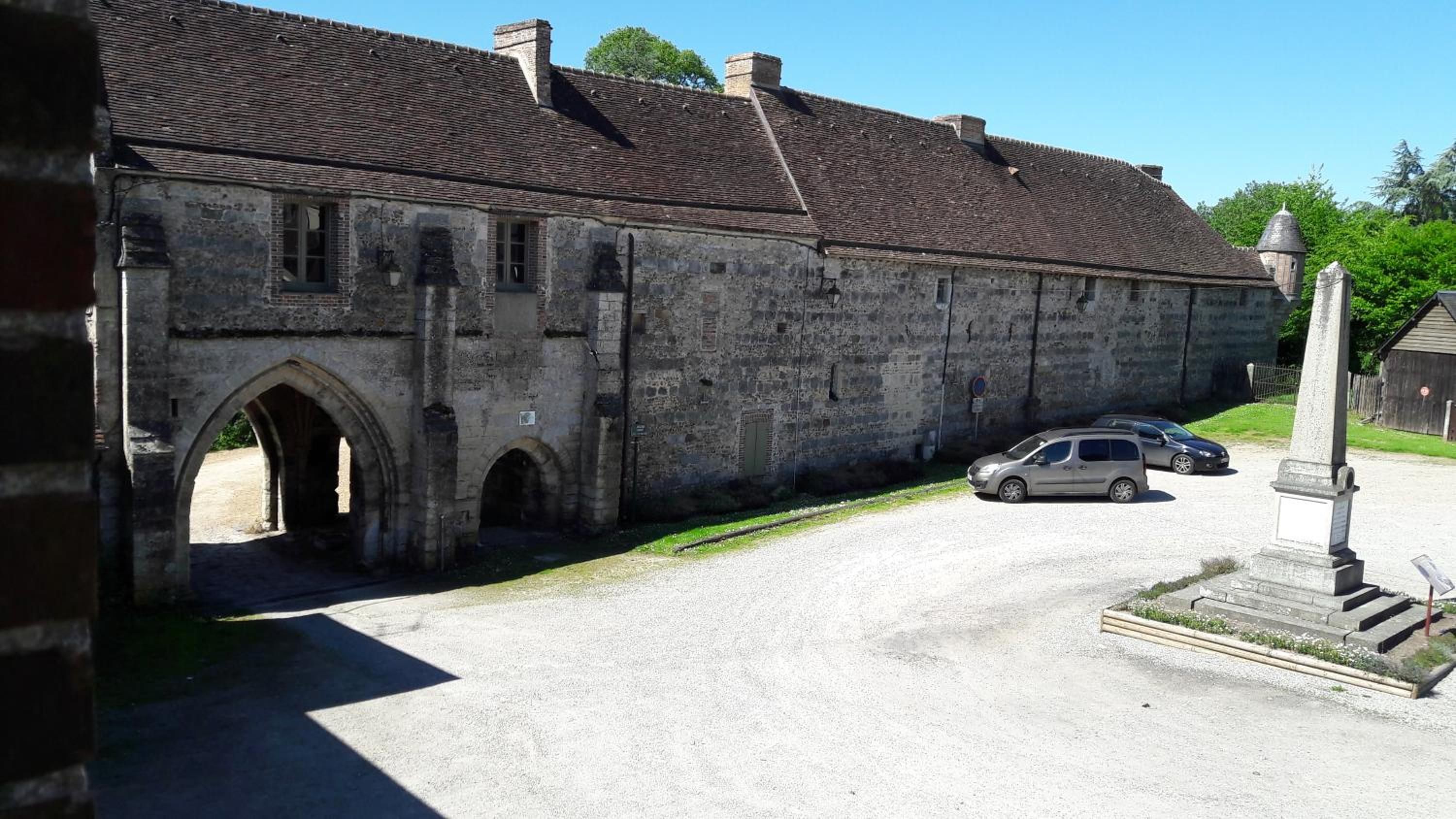A historic stone building with a tiled roof, an arched entrance, and a small parking area with cars.