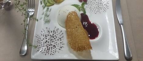 Un plato blanco cuadrado con un alimento frito, una cucharada de crema y salsa de frutos rojos.