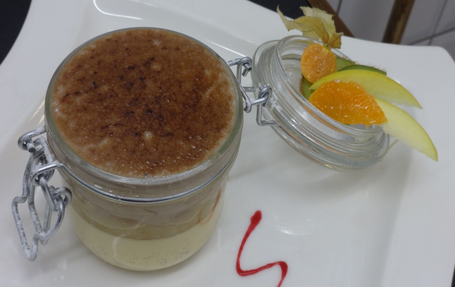 A dessert in a jar with a metal clasp, a small glass jar with fruit and a red sauce on a white plate.