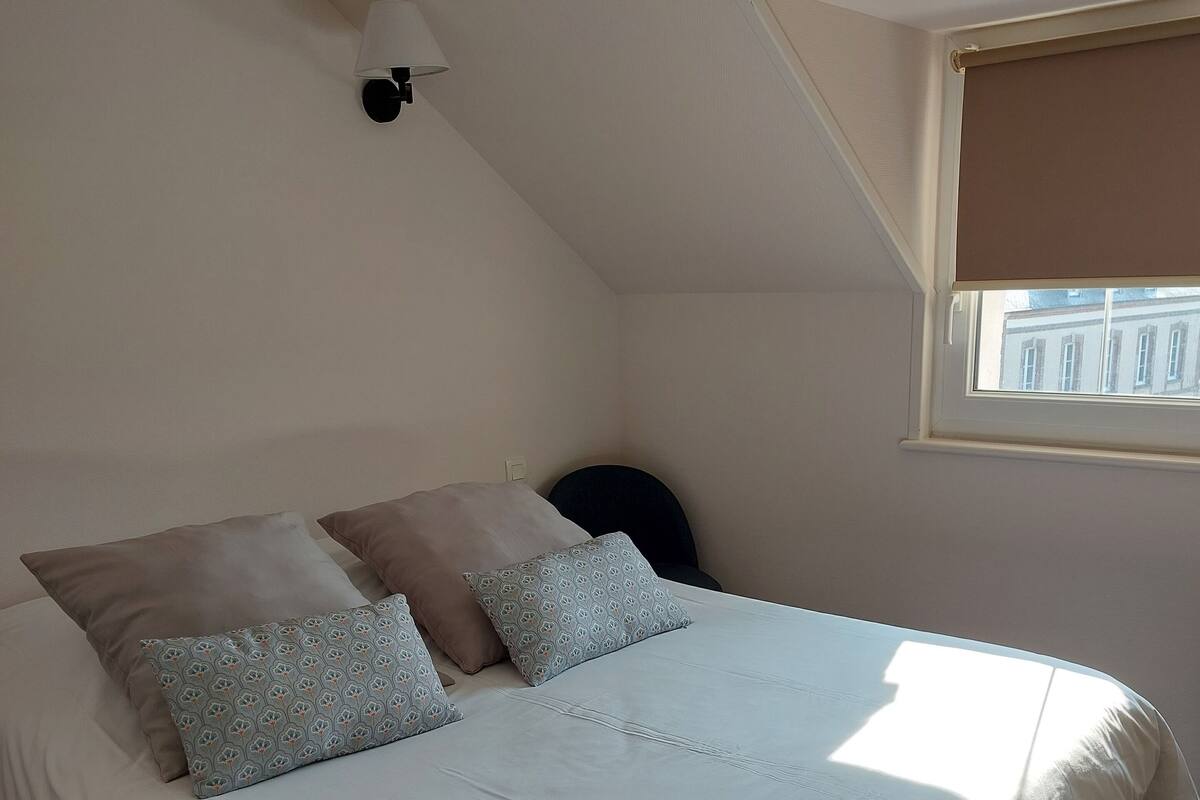 Une chambre à coucher avec un lit, des oreillers, une lampe, une fenêtre et une vue sur les bâtiments à l’extérieur.