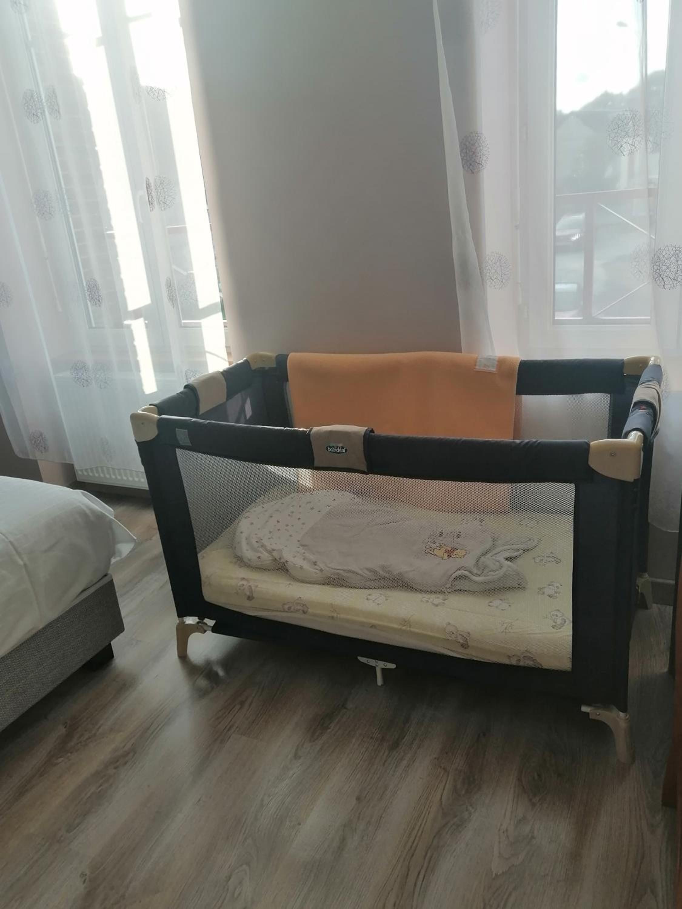 Una cuna para bebé con un techo de malla y un colchón con un diseño de personaje de dibujos animados, ubicada en una habitación con piso de madera y una cama al fondo.