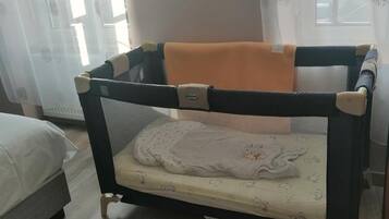 Una cuna para bebé con un techo de malla y un colchón con un diseño de personaje de dibujos animados, ubicada en una habitación con piso de madera y una cama al fondo.