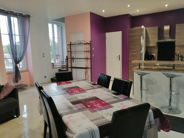 Un comedor con una mesa dispuesta para cuatro, una cocina con electrodomésticos modernos y un ventanal con cortinas.