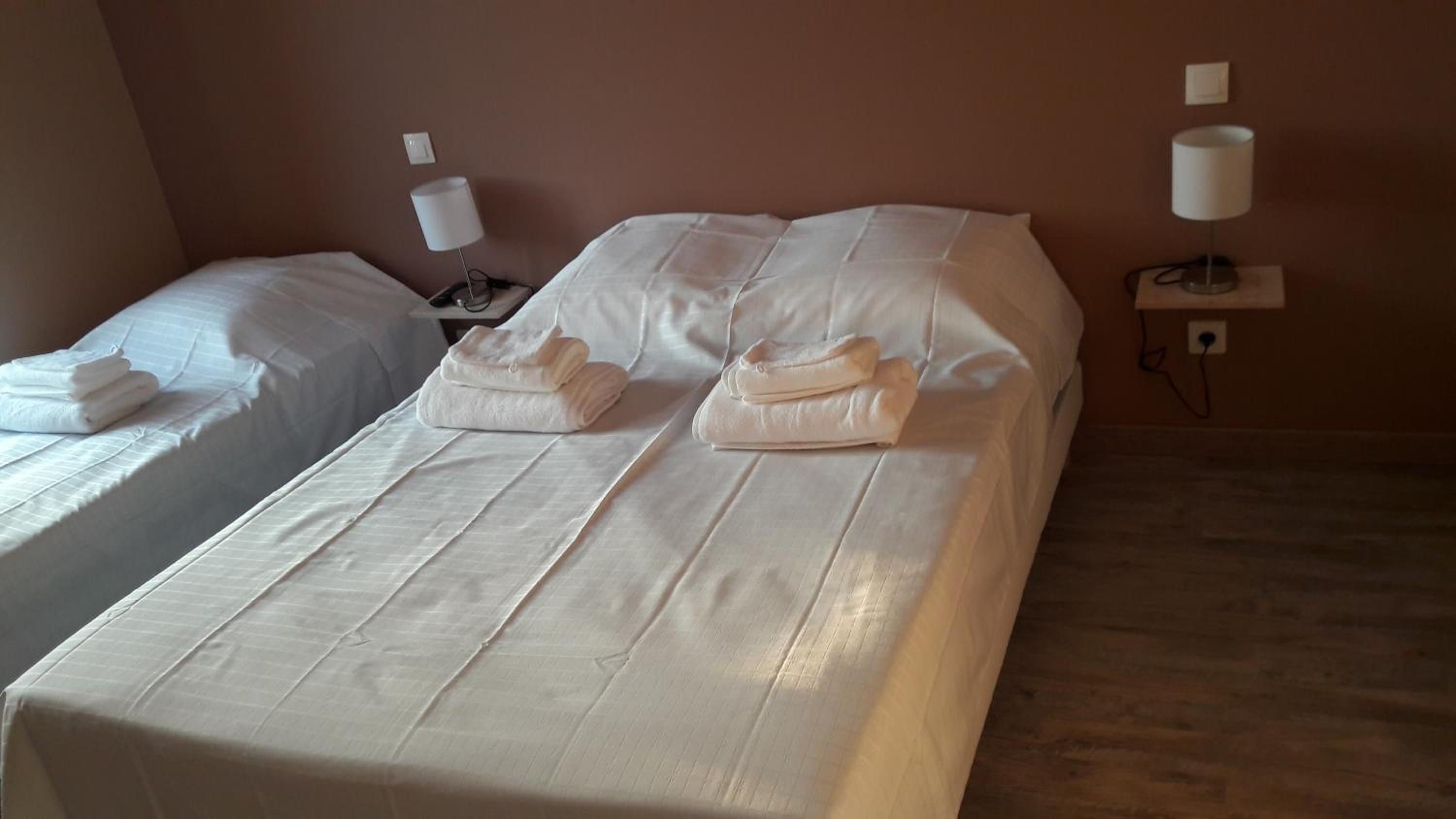 Habitación de hotel con dos camas individuales, cada una con una colcha y almohadas blancas, y una mesita de noche con una lámpara.