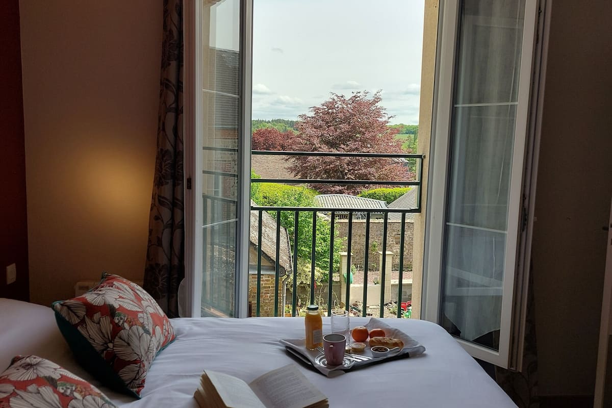 Une chambre d’hôtel avec un lit, une fenêtre ouverte et un balcon donnant sur un paysage verdoyant et un arbre.