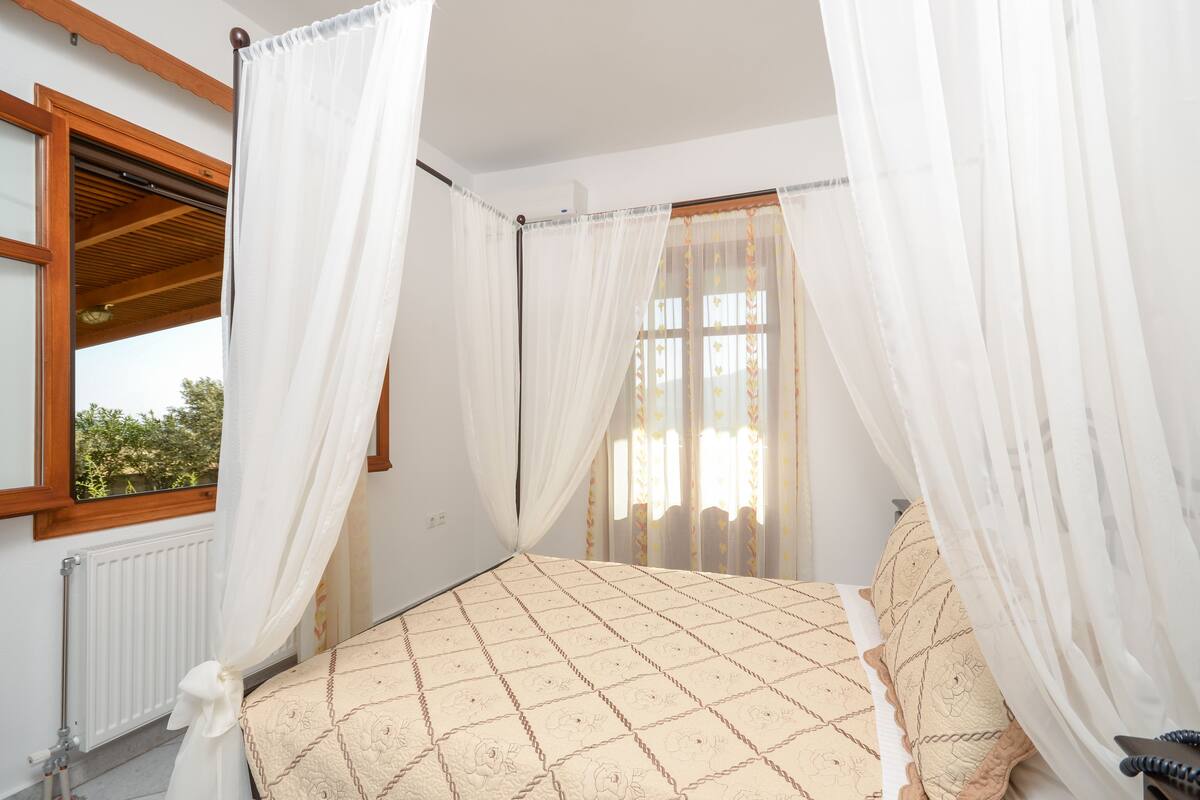 A bedroom with a bed, white curtains, a window with a view of greenery, and a radiator.