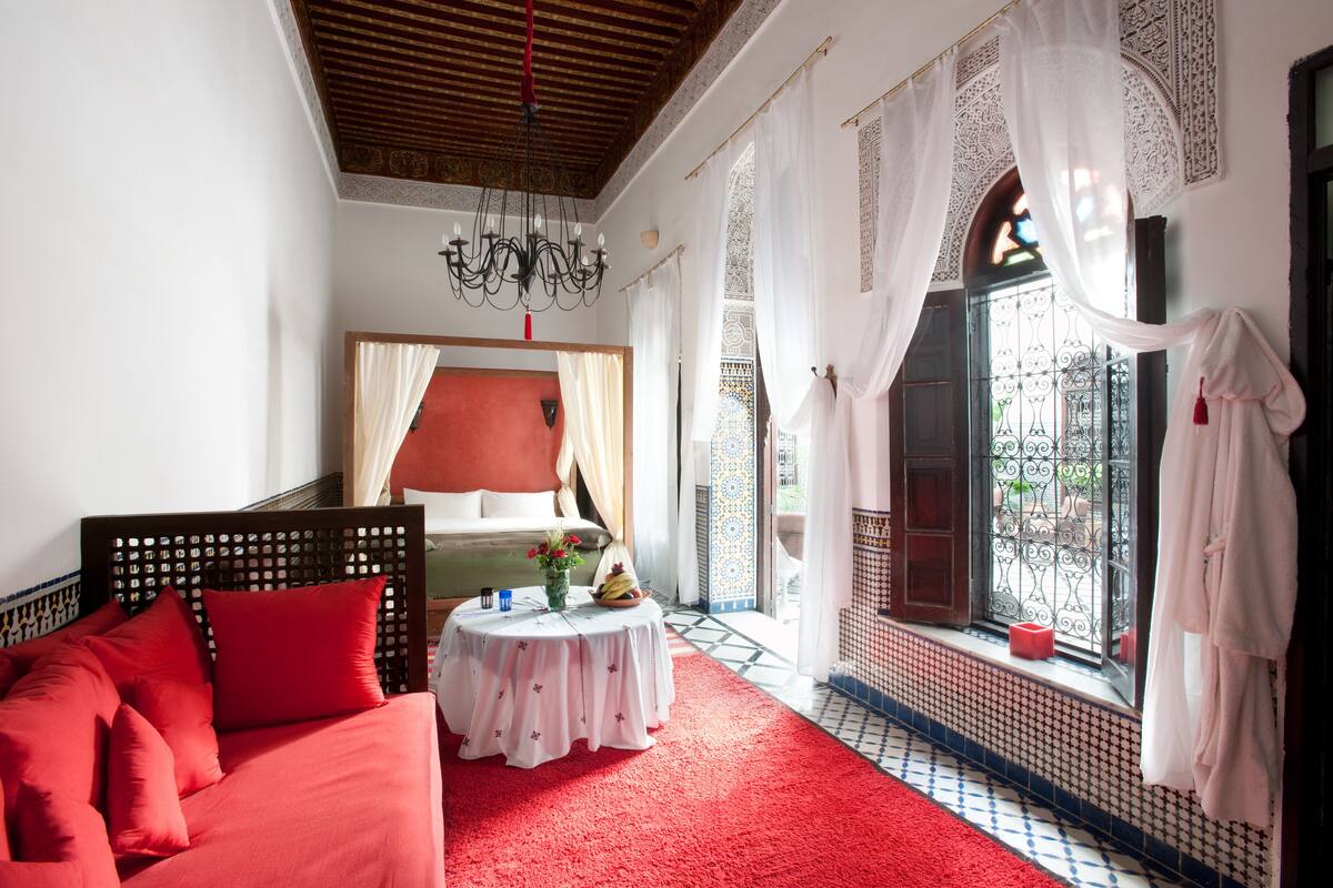 A bedroom with a red sofa, a bed with a red headboard, a small round table with a vase and fruit, and a window with white curtains and a view of the outside.