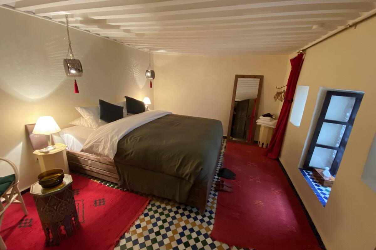 A bedroom with a bed, a nightstand, a lamp, a red and white checkered floor, a window, and a door leading to another room.