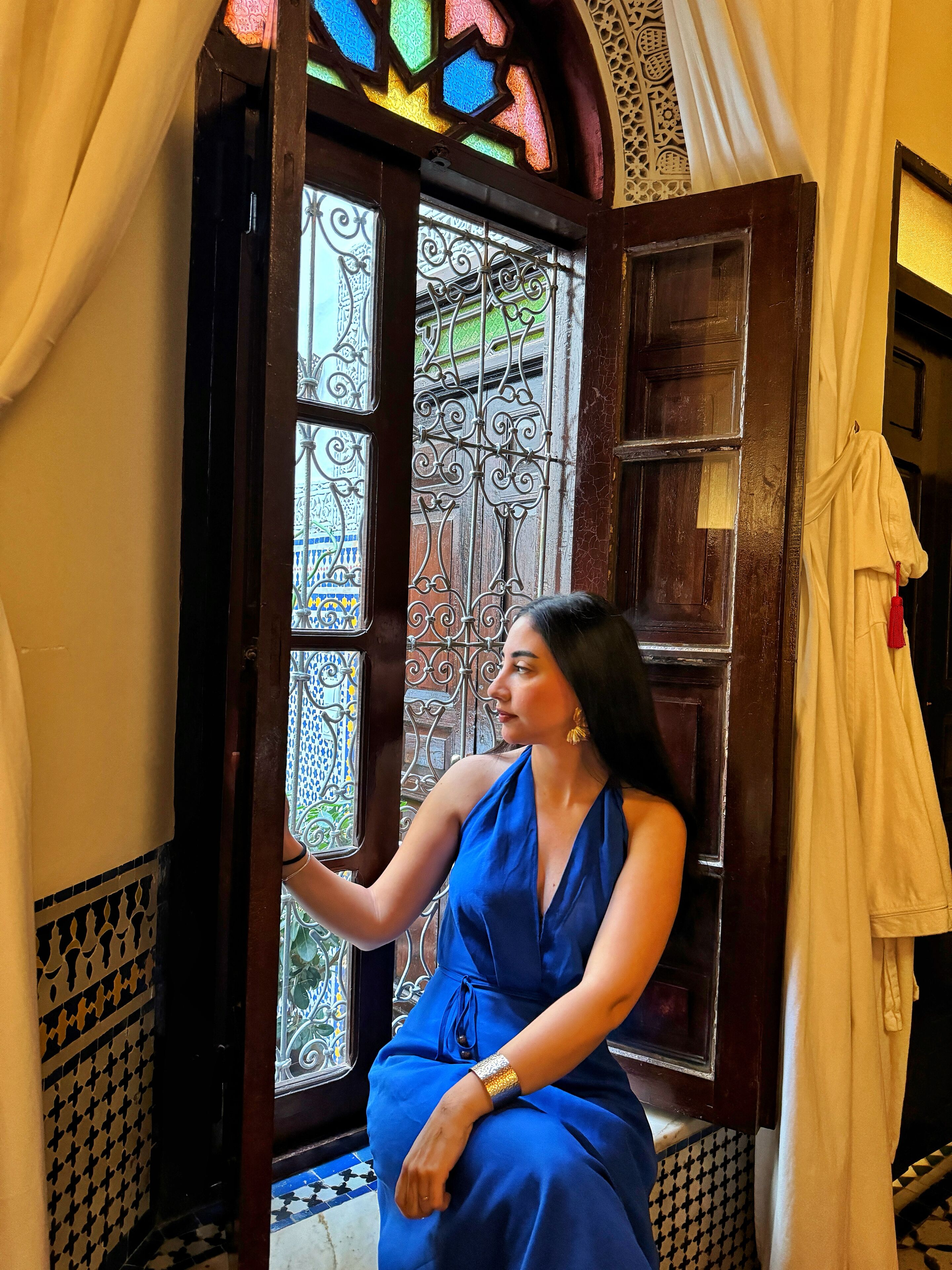 Una mujer sentada junto a una ventana con barrotes metálicos ornamentados y una puerta de madera.