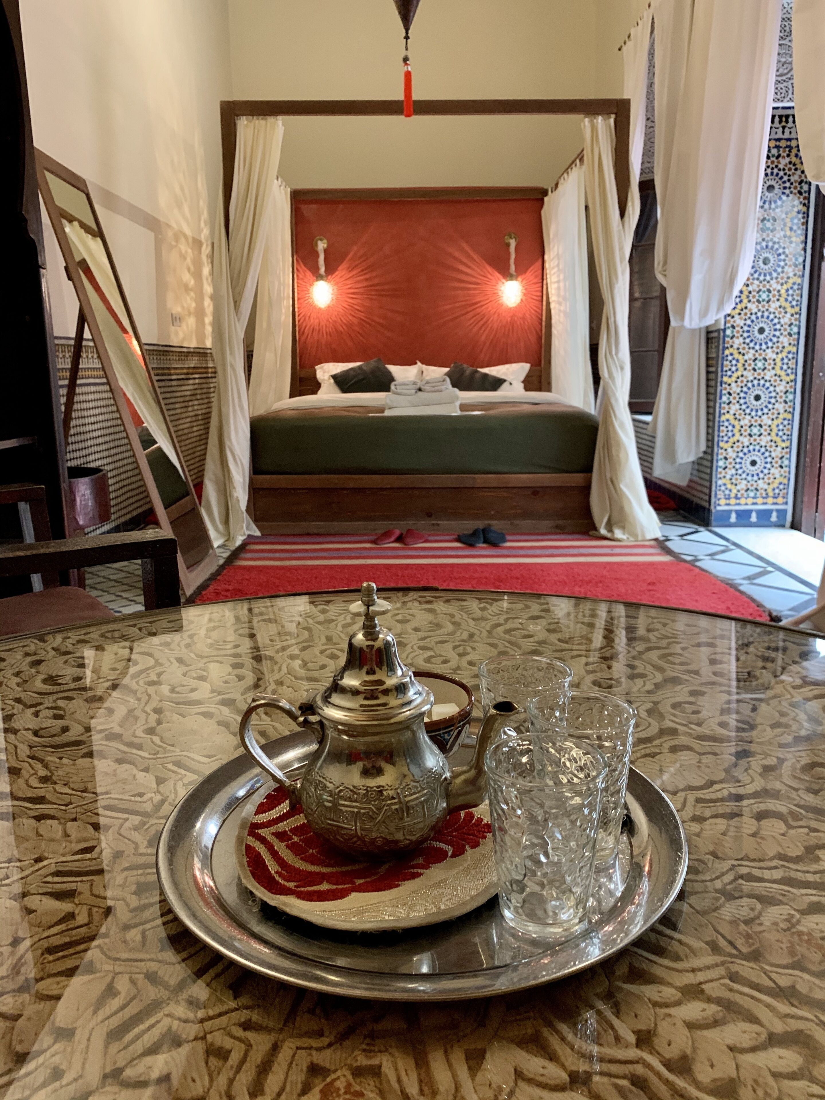A bedroom with a bed, a bedside table with a teapot and glasses, and a red carpet.