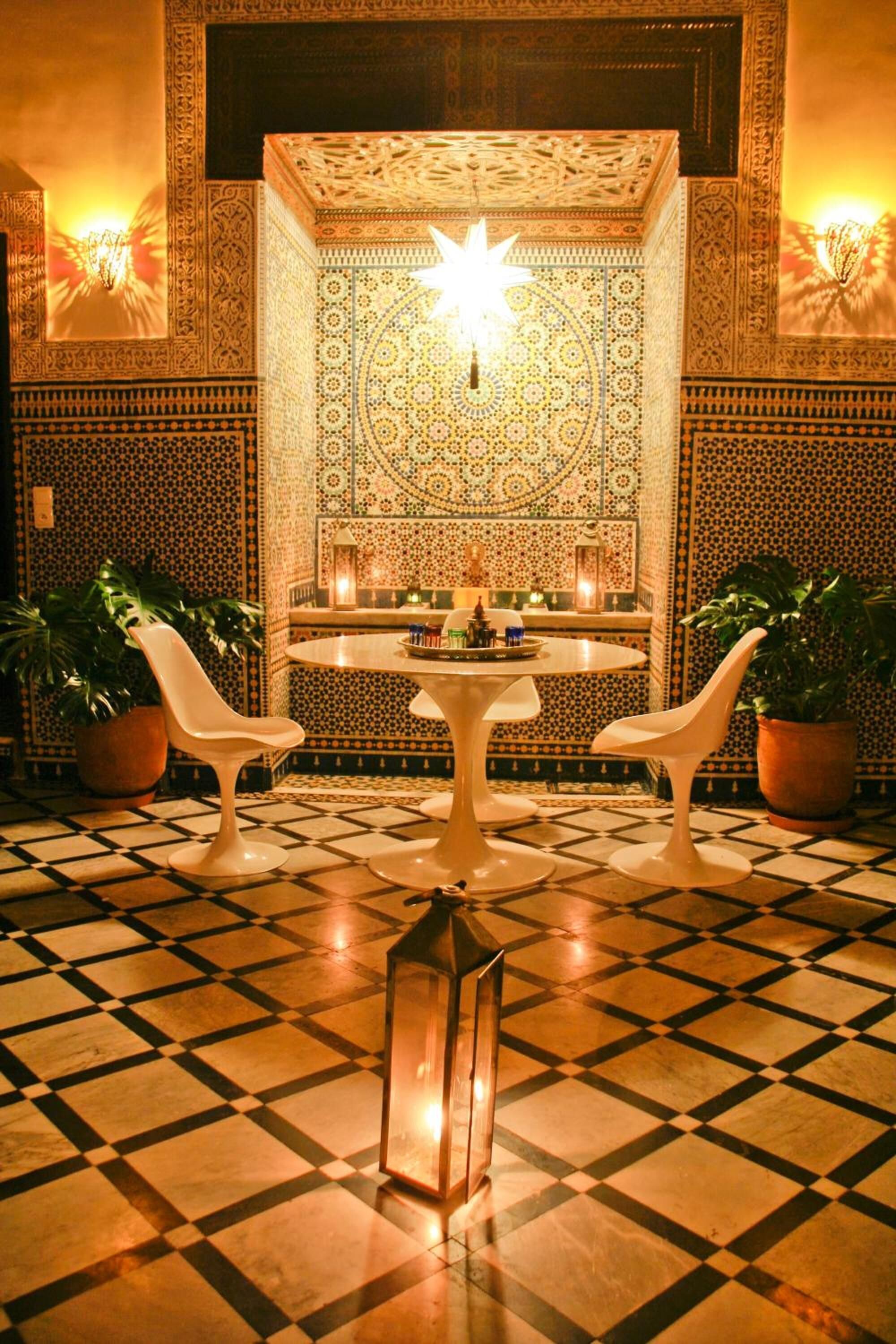 Un interior al estilo marroquí con intrincados diseños de azulejos, una mesa central con sillas y plantas en macetas.