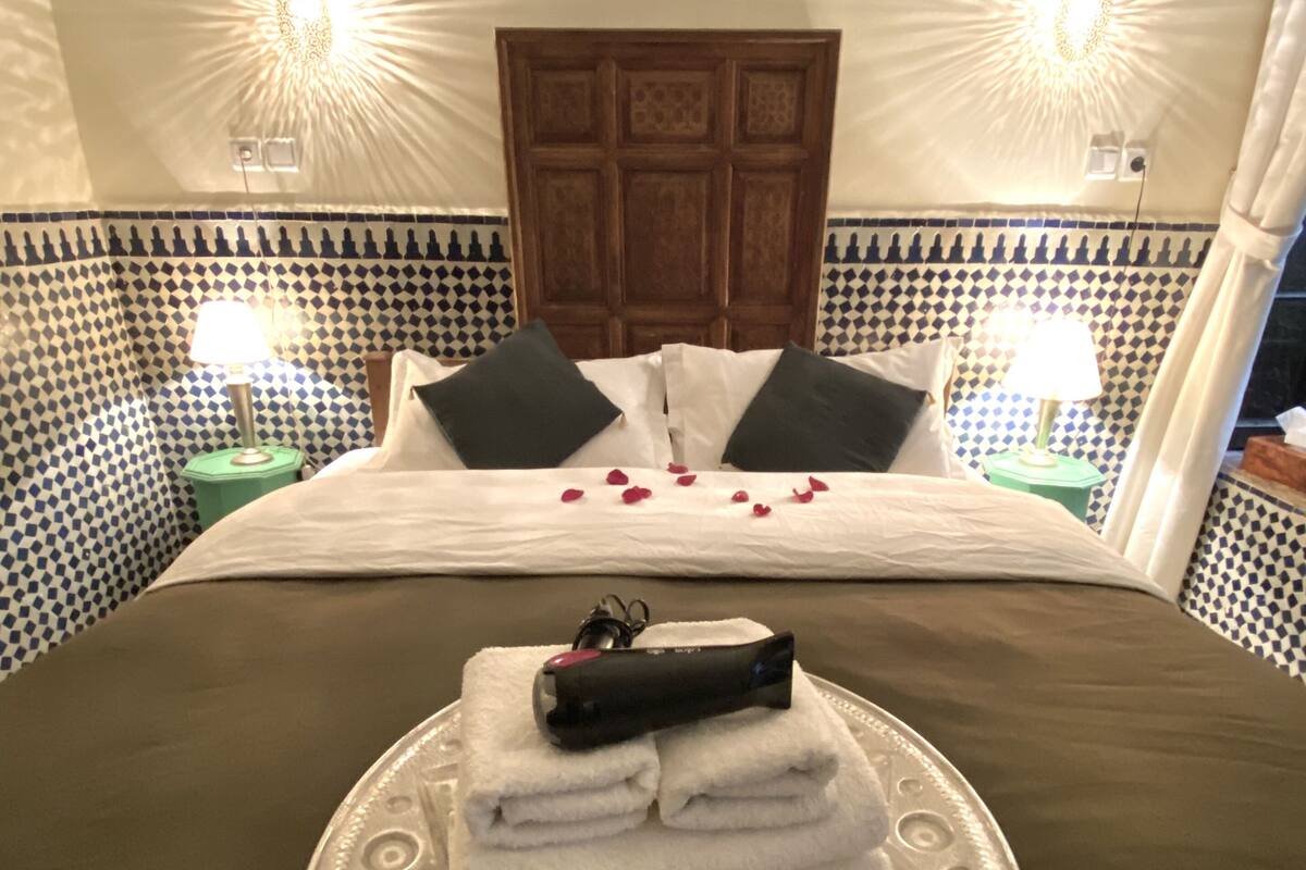 A neatly made bed with a towel stack, rose petals, and a remote control on a tray.
