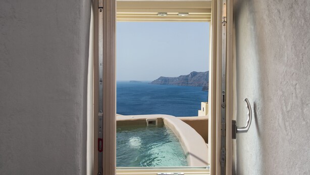 A view from a room with a large window overlooking a pool and the sea. - Ammos Oia Mansion (Santorini)
