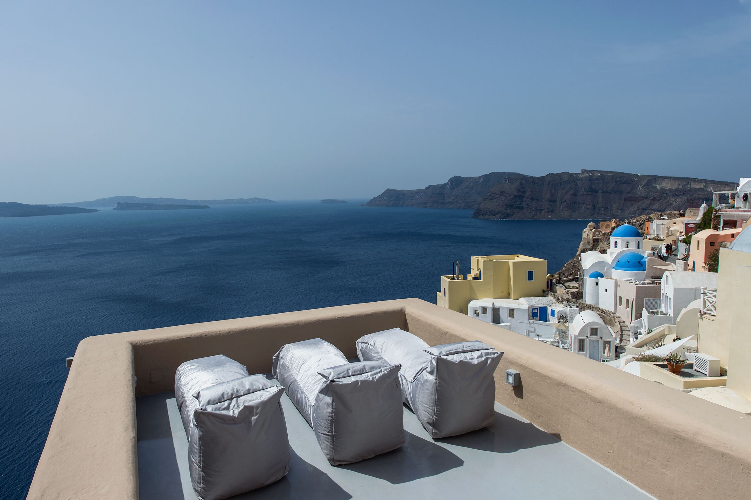 A balcony with white cushioned seating overlooking a coastal town with blue-domed buildings and a view of the sea.