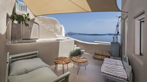 A balcony with a bed, two round tables, a striped cushion, and a view of the sea. - Ammos Oia Mansion (Santorini)