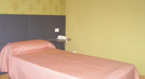 Una cama individual con edredón rosa en una habitación con pared lisa y una mesita de noche pequeña con teléfono.