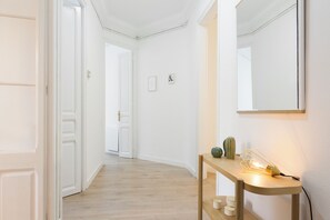 Um corredor com paredes brancas, piso de madeira, um espelho, uma mesa com abajur e uma porta.