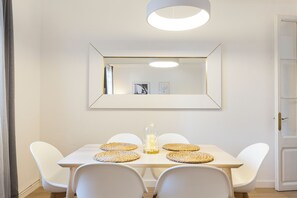Uma sala de jantar com mesa branca, quatro cadeiras brancas, uma vela central e um espelho grande na parede.
