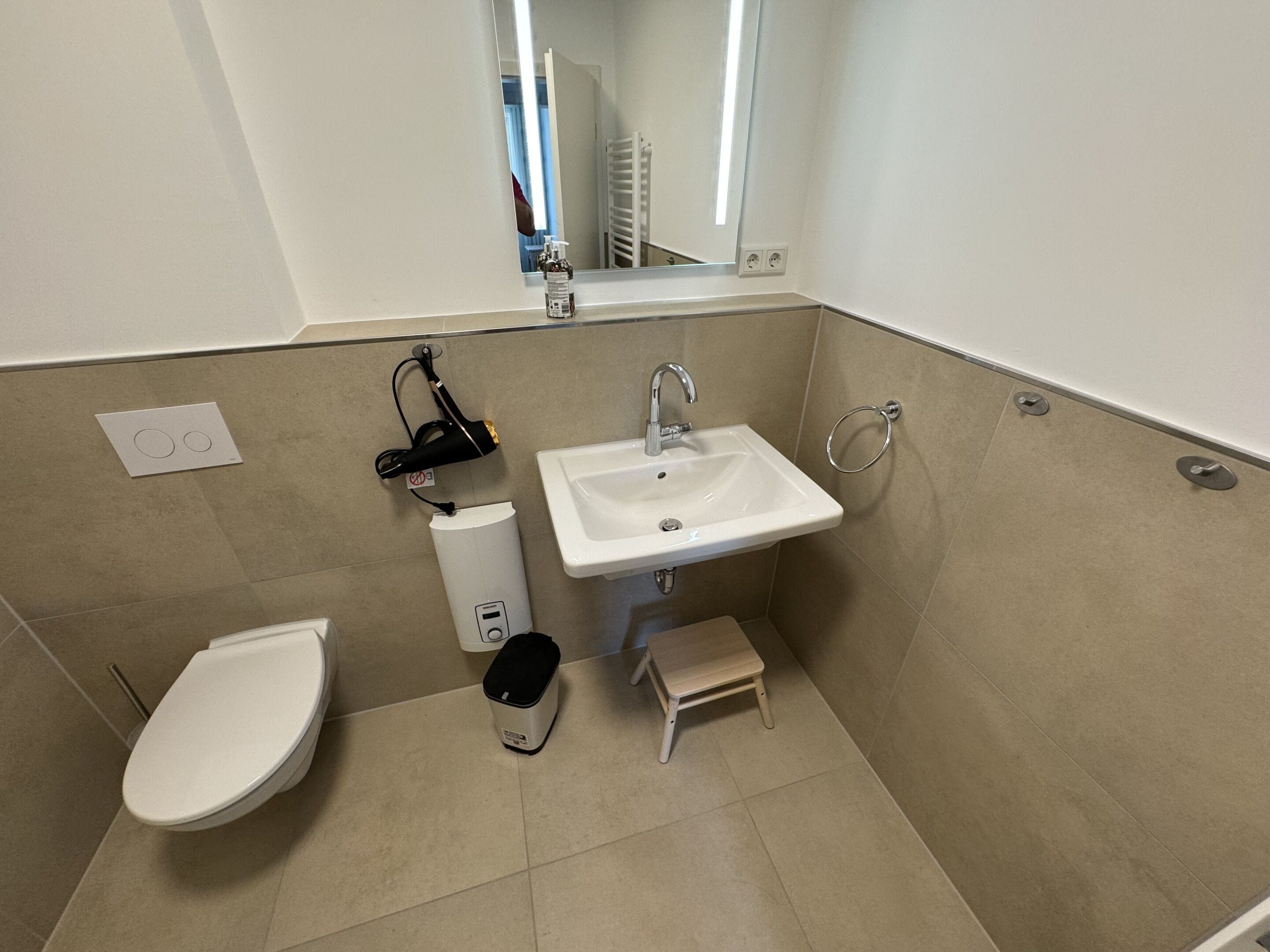 Un baño con un lavamanos blanco, un inodoro, un taburete pequeño y un secador de pelo sobre un soporte en la pared.