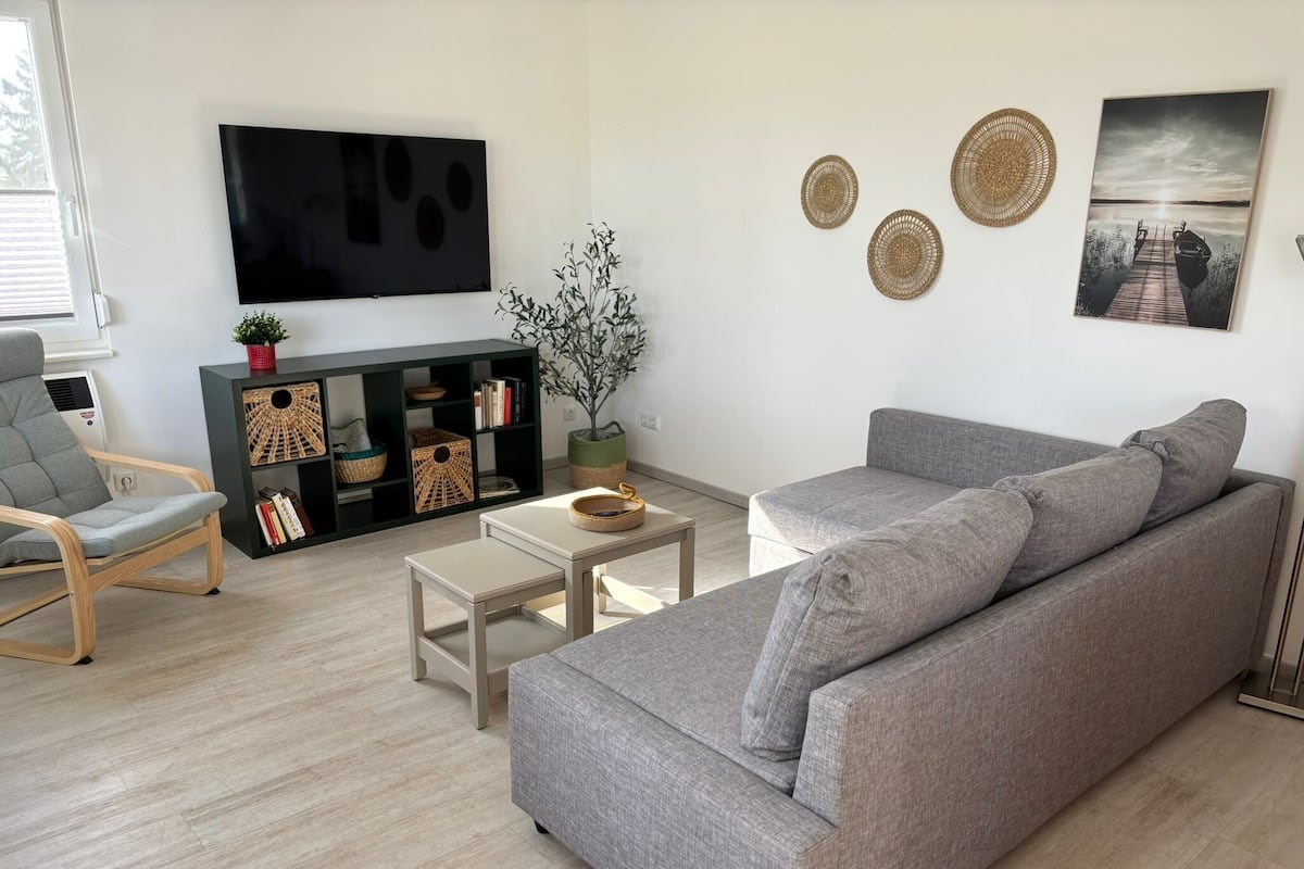 A living room with a grey sofa, a wooden chair, a small table, a black entertainment unit with woven baskets, and a flat-screen TV.