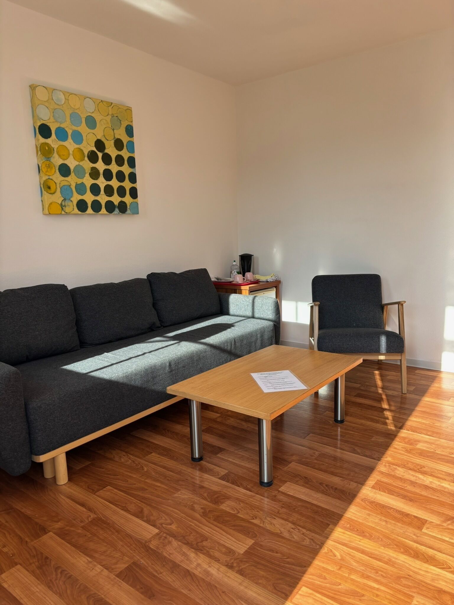 Una sala de estar moderna con un sofá gris, una mesa de centro de madera y una silla. Hay una mesita con una lámpara y una taza encima. Un cuadro abstracto y colorido adorna la pared.