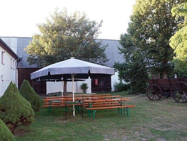 An outdoor seating area with picnic tables and a large umbrella, surrounded by trees and a building with a metal roof. - Restaurant und Pension Zur Wildgans (Arendsee)