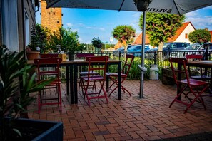 Un patio al aire libre con sillas y mesas rojas, suelo de ladrillo y vista a un edificio de ladrillo y árboles al fondo.