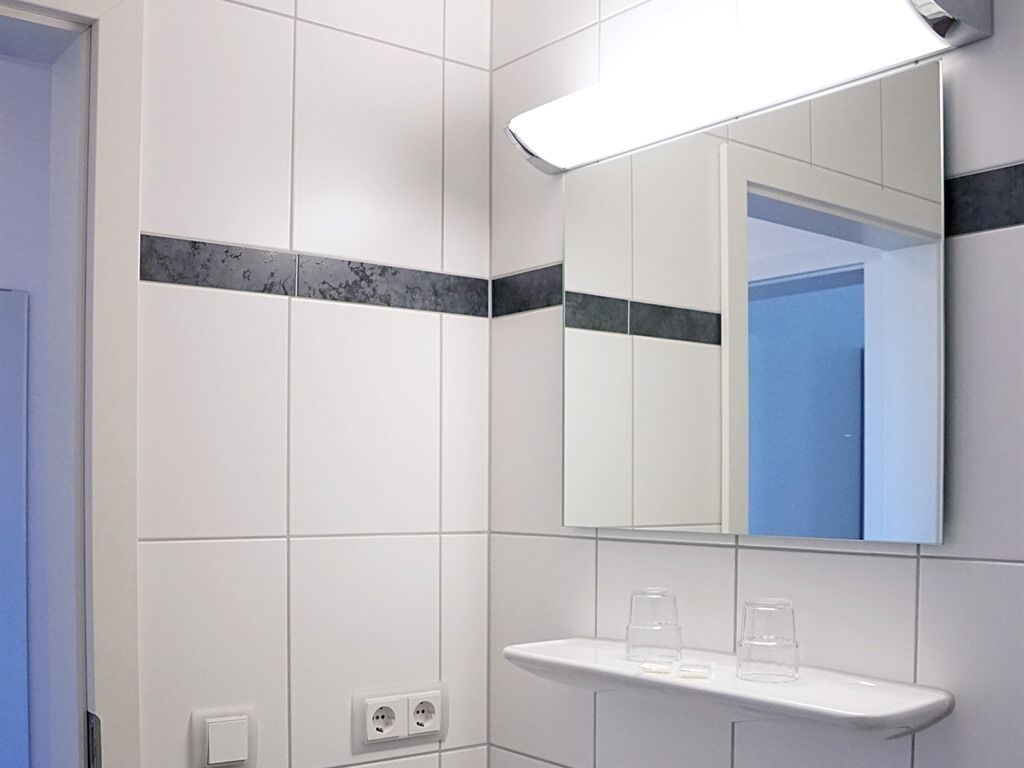 A bathroom with white tiles, a mirror, and a shelf with glasses.
