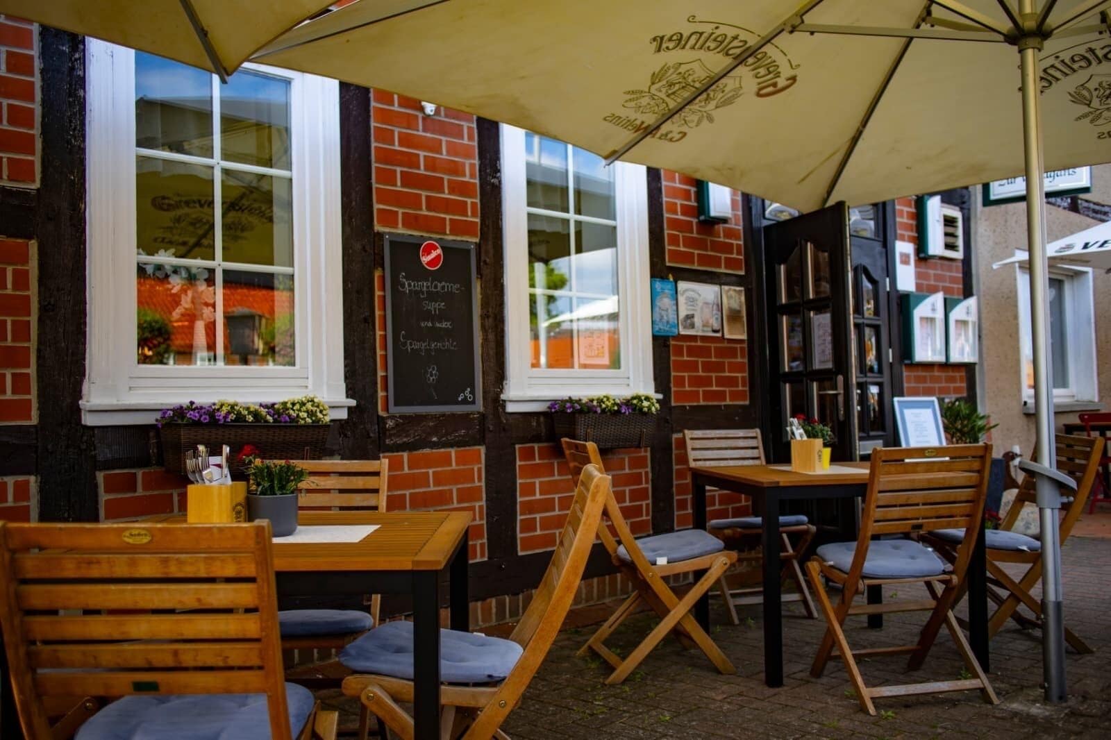 An outdoor café with wooden tables and chairs, a chalkboard menu, and a brick wall.