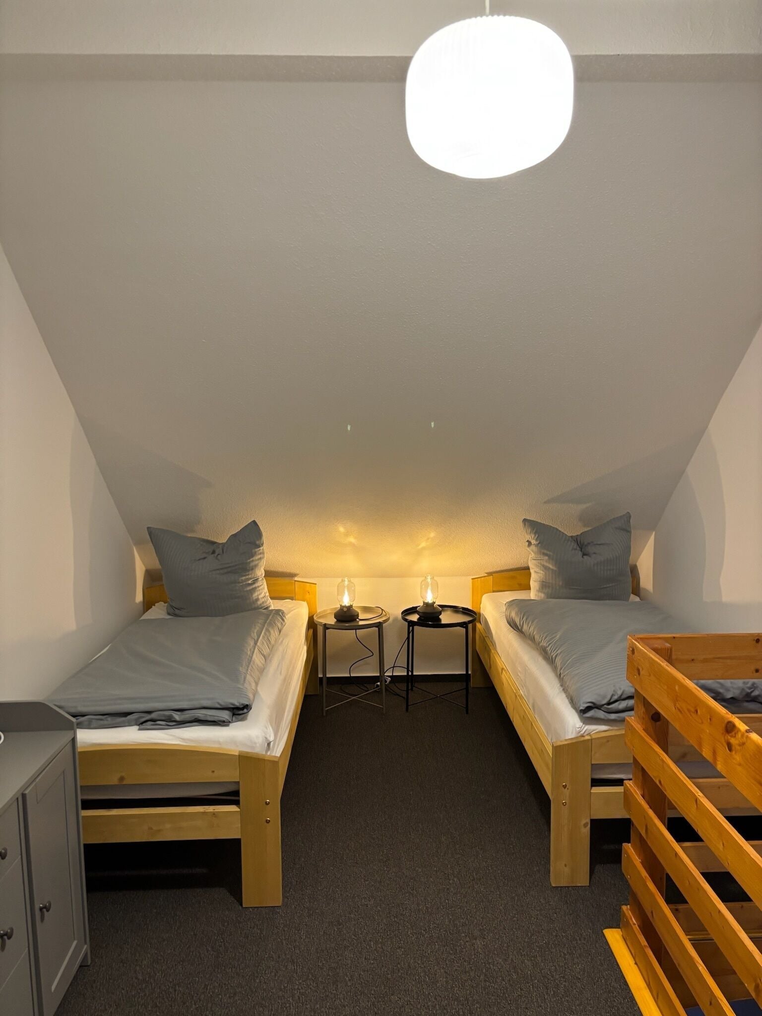 A small, cozy room with two single beds, a wooden bench, a desk with a chair, and a hanging light fixture.