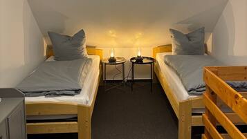 A small, cozy room with two single beds, a wooden bench, a desk with a chair, and a hanging light fixture.