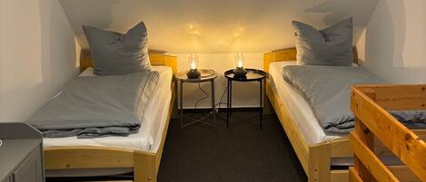 A small, cozy room with two single beds, a wooden bench, a desk with a chair, and a hanging light fixture.