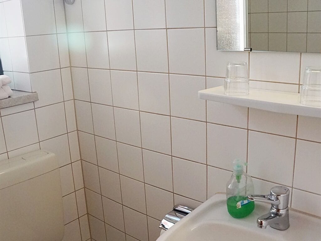 Un baño con azulejos blancos, un lavamanos con una botella verde y un espejo sobre él.