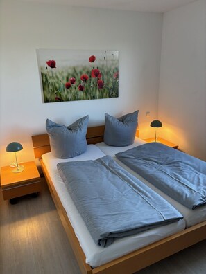 Un dormitorio con una cama de madera, dos mesitas de noche, una lámpara en cada una y un cuadro colgado en la pared con flores rojas.