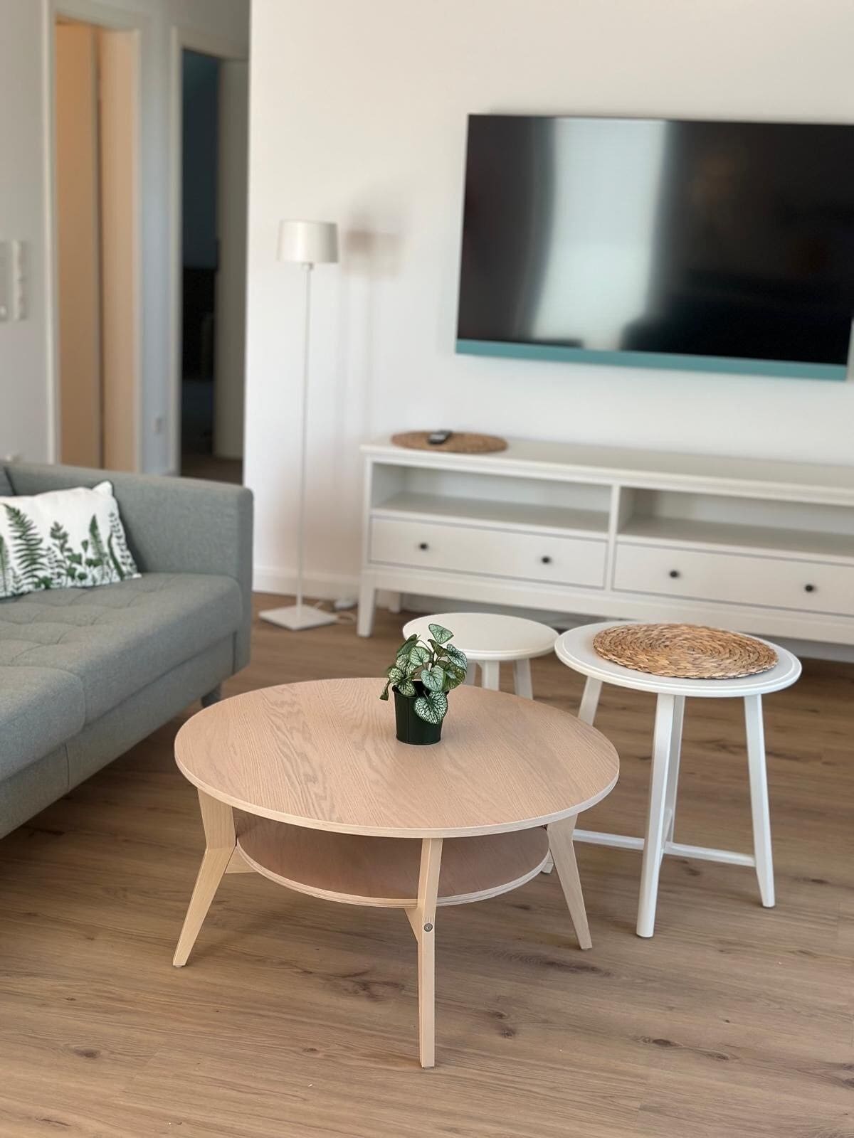 Una sala de estar moderna con un sofá gris, una mesa de centro redonda de madera, una pequeña planta en maceta, un mueble de televisión blanco y un televisor de pantalla plana.