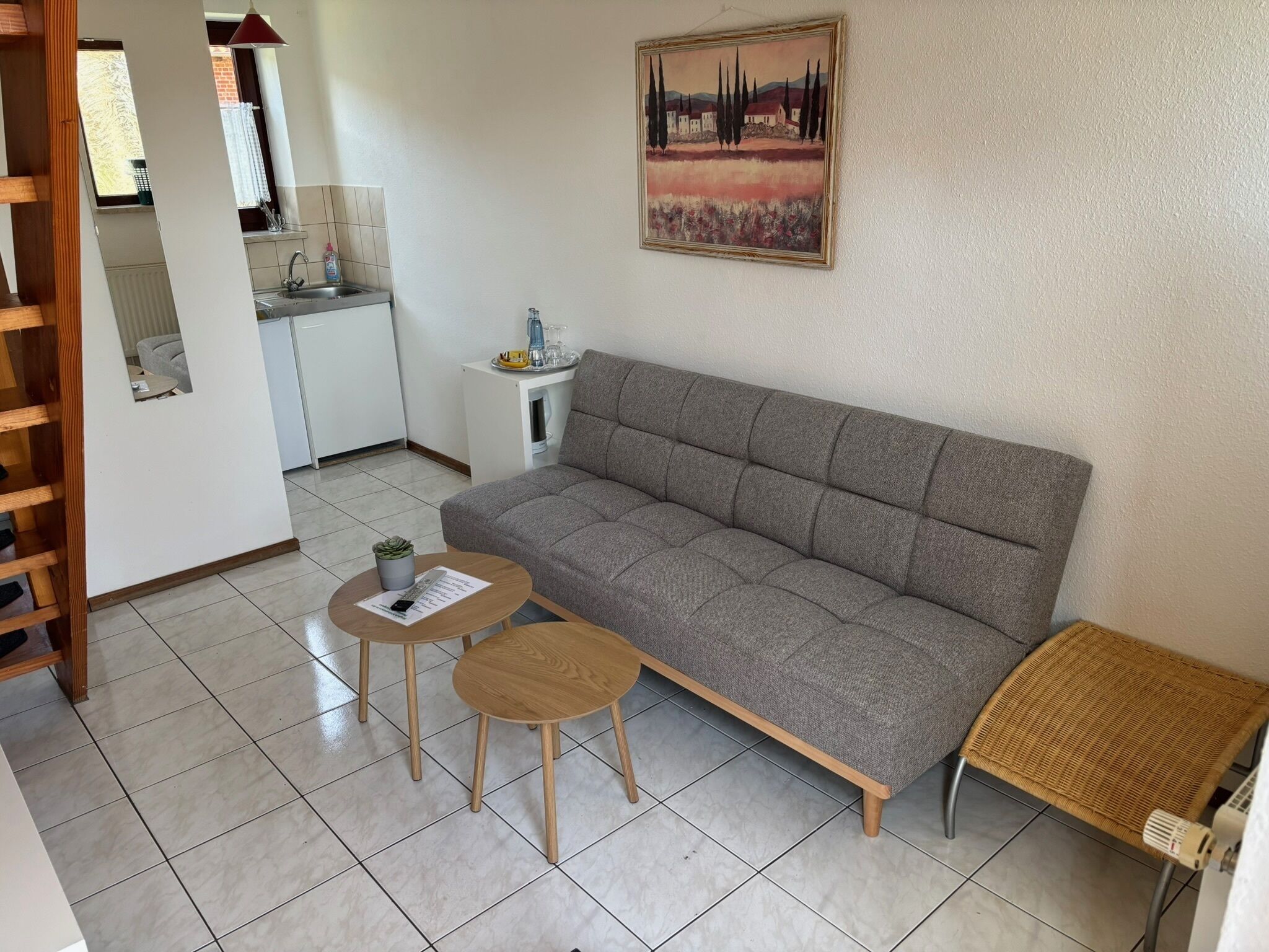 Sala de estar con un sofá gris, dos mesas de madera, un banco de mimbre, una zona de cocina con un lavabo y una ventana, y un cuadro en la pared.