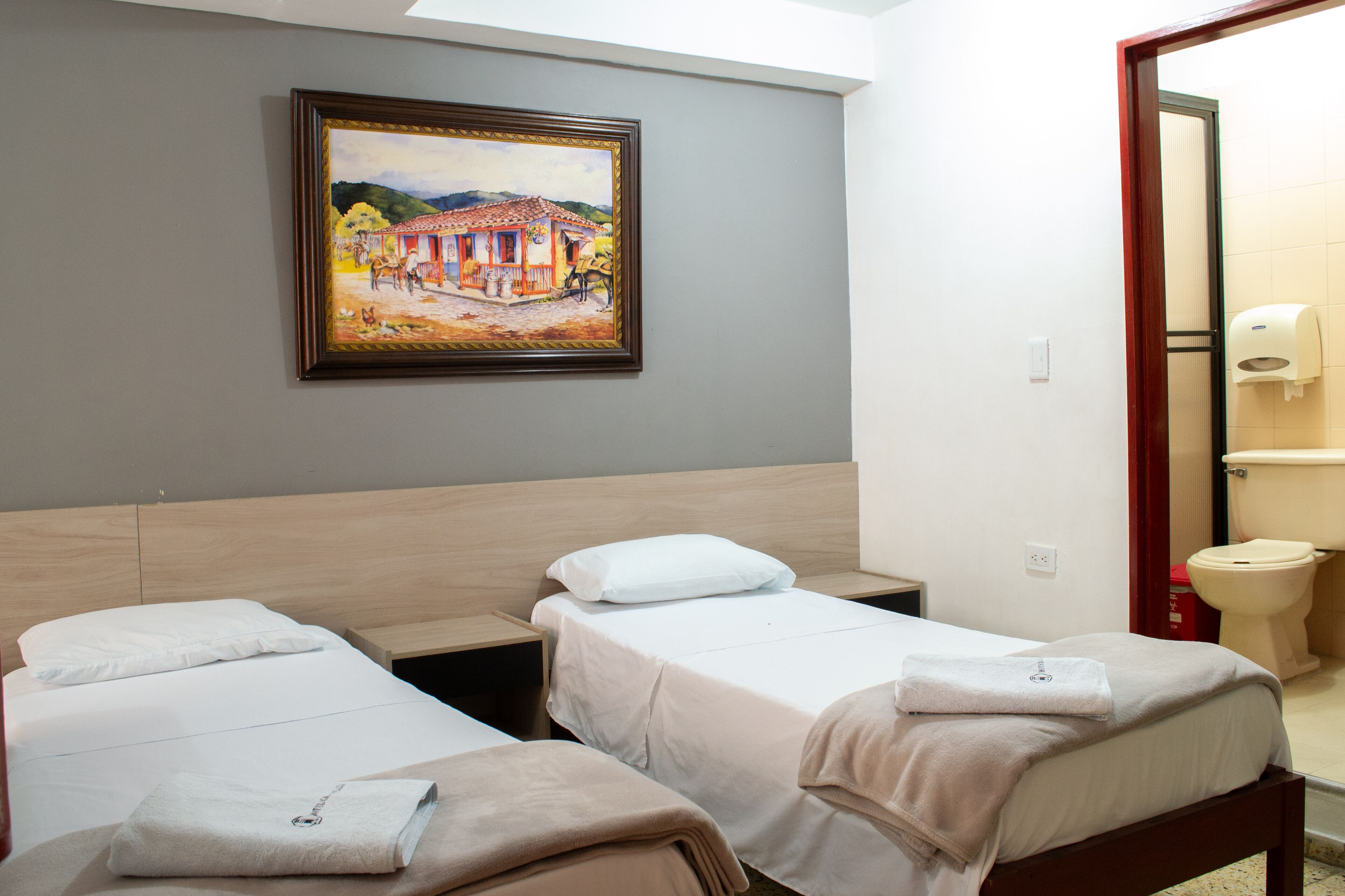Habitación de hotel con dos camas individuales, un cuadro en la pared y un baño visible a través de una puerta abierta.