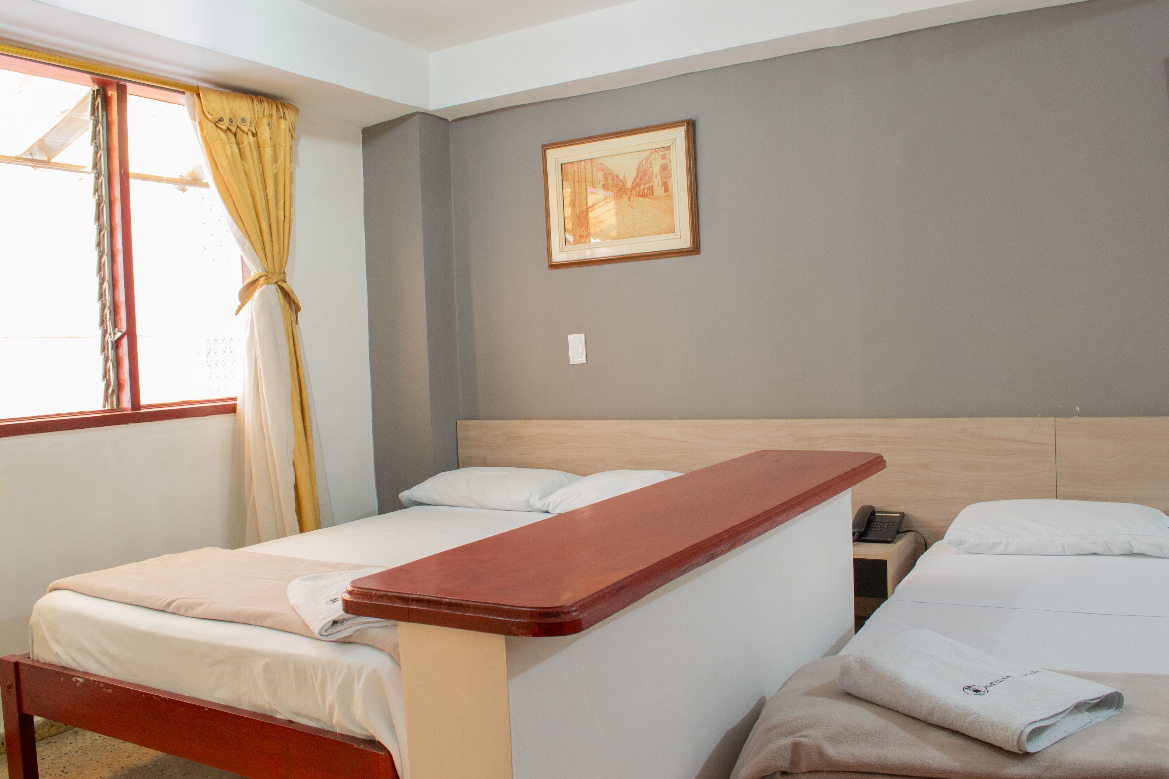 A hotel room with two single beds, a wooden headboard, a small bedside table with a phone, and a window with curtains.
