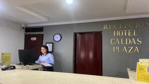 Mostrador de recepción en el Hotel Caldas Plaza, con un recepcionista, un reloj y un cartel que indica las horas de entrada y salida.