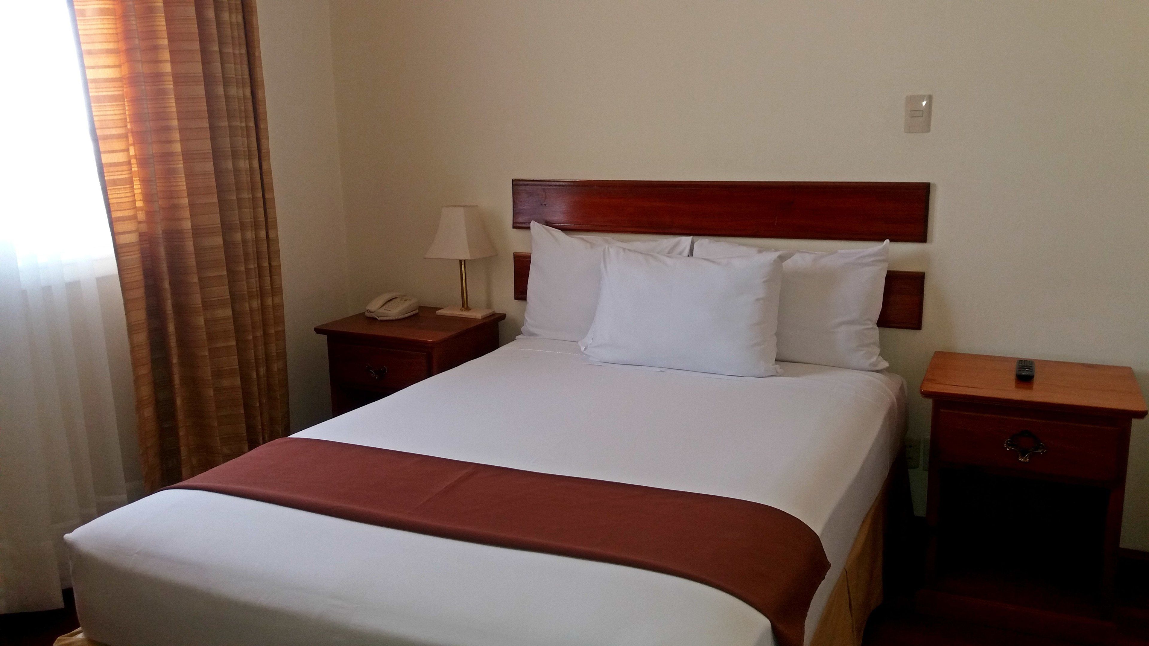 A hotel room with a single bed, a nightstand with a phone and lamp, a wooden headboard, and a small drawer with a remote control.